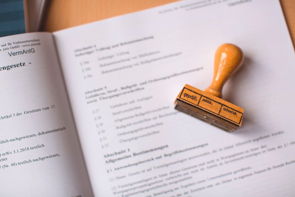 A close-up image showing an open document or legal paper with typed text, partially blurred, laid out on a flat surface. Resting on the paper is a rubber stamp with a wooden handle, stamped with black