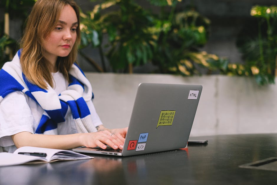 A young woman with shoulder-length light brown hair, wearing a white shirt and a blue and white striped scarf, is sitting at a dark table outdoors, working on a silver laptop decorated with small colo