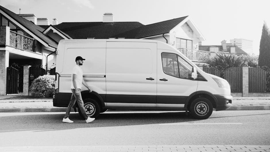 A man and a woman are preparing boxes for a home relocation inside the open rear of a white moving van parked outside a residential property. The man, wearing a white polo shirt, blue jeans, and a dar