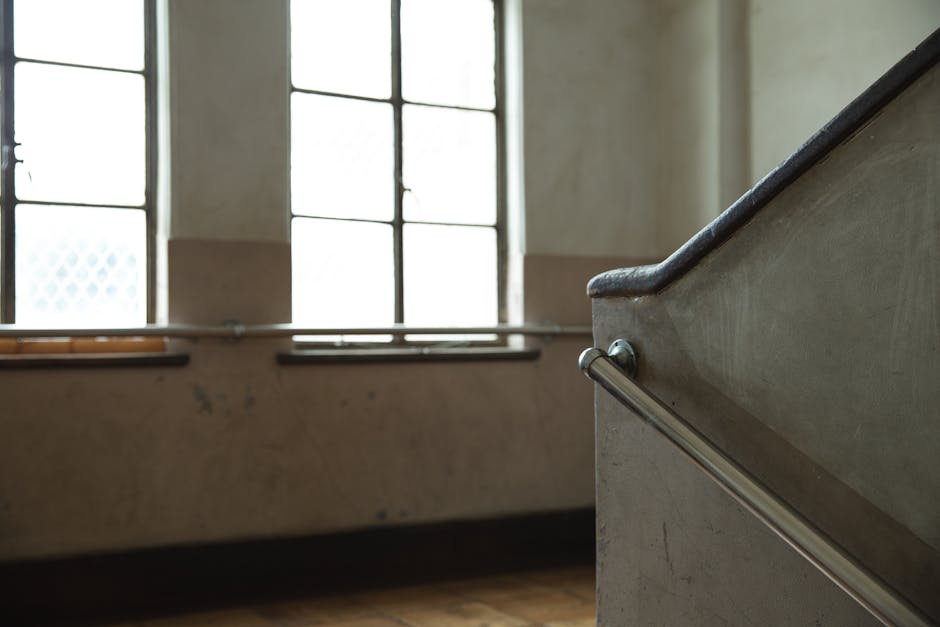 A top-down view of a spiral staircase inside a building, showing multiple landings and flights of stairs with beige tiled flooring. The staircase has black metal railings and a wooden handrail, with t
