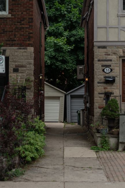 A narrow residential driveway leading up to a garage beneath a balcony of a white and beige multi-storey building with external stairs and black metal railings. The driveway is paved with concrete, sh